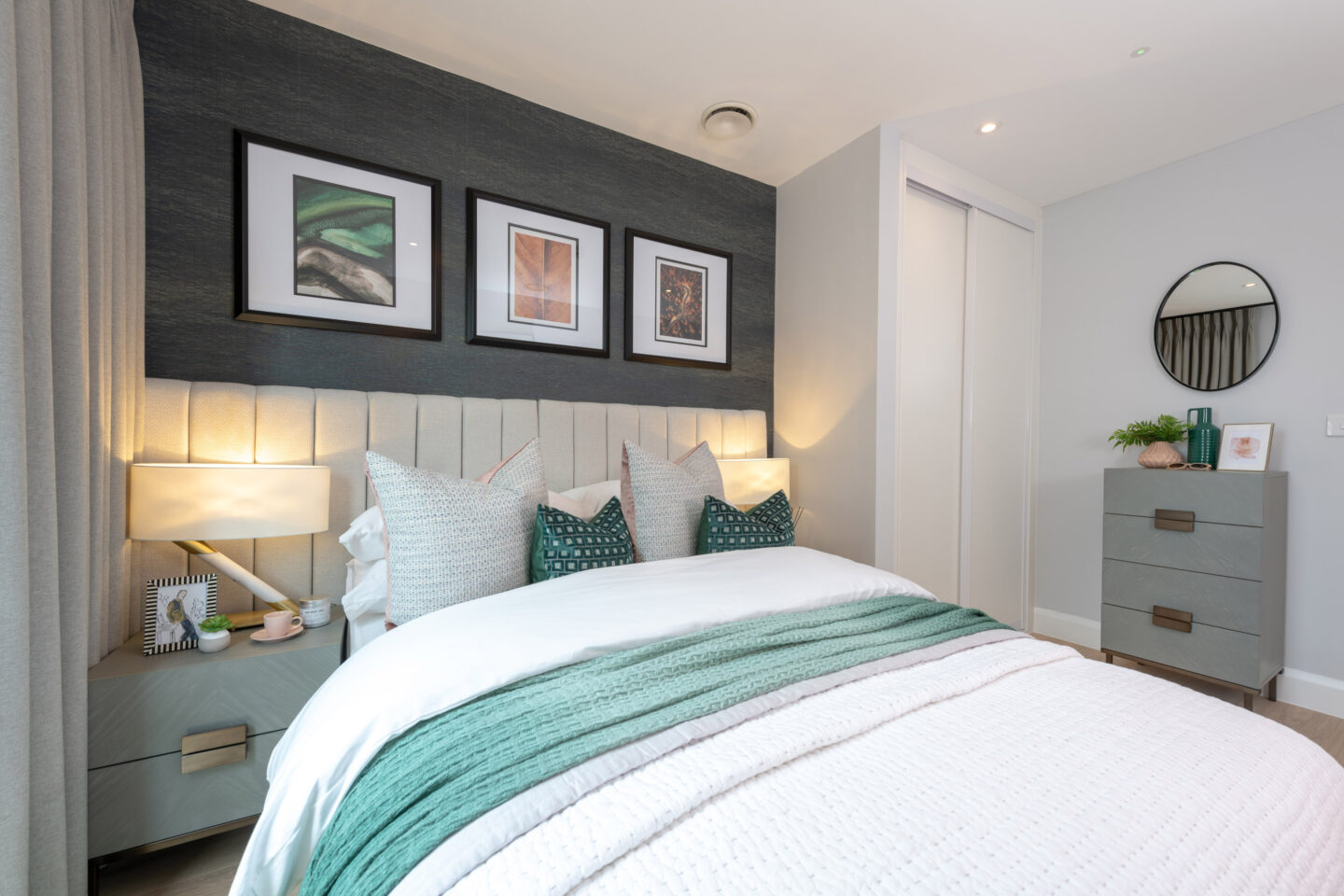 Modern bedroom with a neatly made bed, green and white bedding, two bedside tables with lamps, three framed pictures above the headboard, and a chest of drawers with a round mirror.