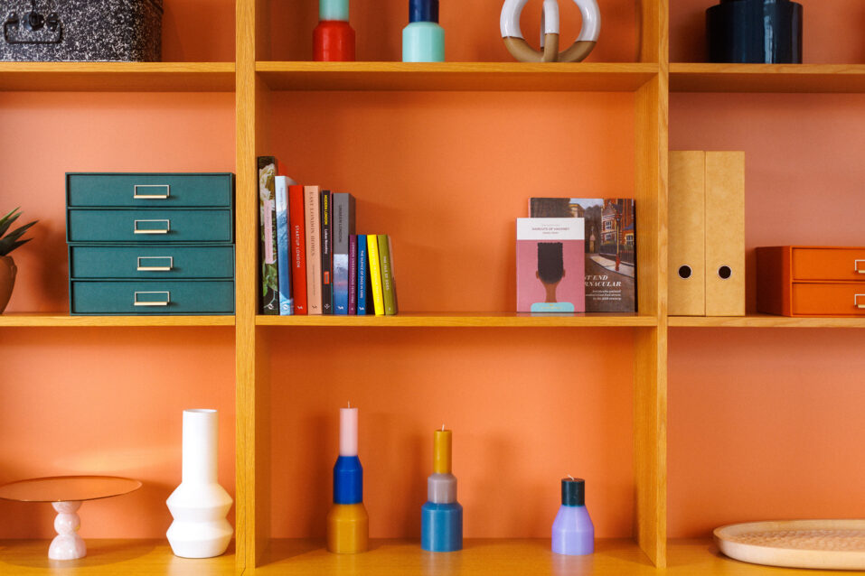 Wooden shelving unit against an orange wall, displaying books, colourful vases, boxes, and decorative objects arranged neatly in compartments.