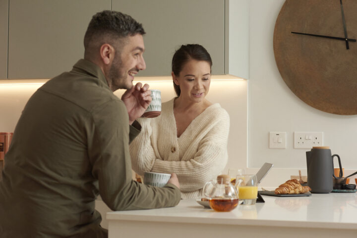 A man and a woman stand in a modern kitchen, holding mugs and talking. Croissants, juice, teapot, and an open tablet are on the worktop. A large clock is on the wall behind them.