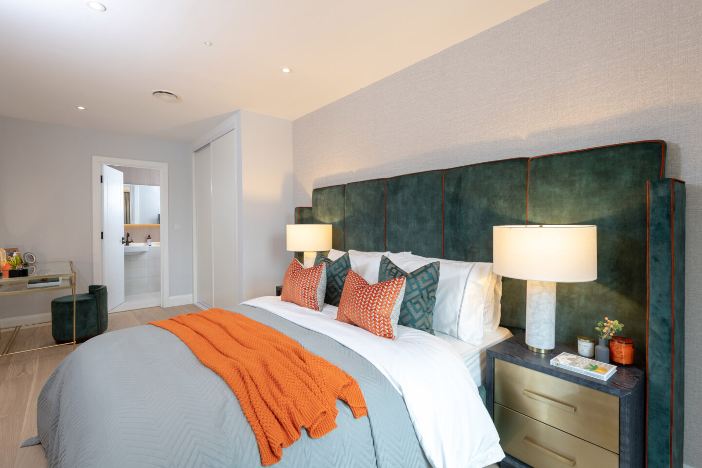 Modern bedroom with a large green upholstered headboard, bedside lamps, orange accents, and an open door leading to a bathroom.