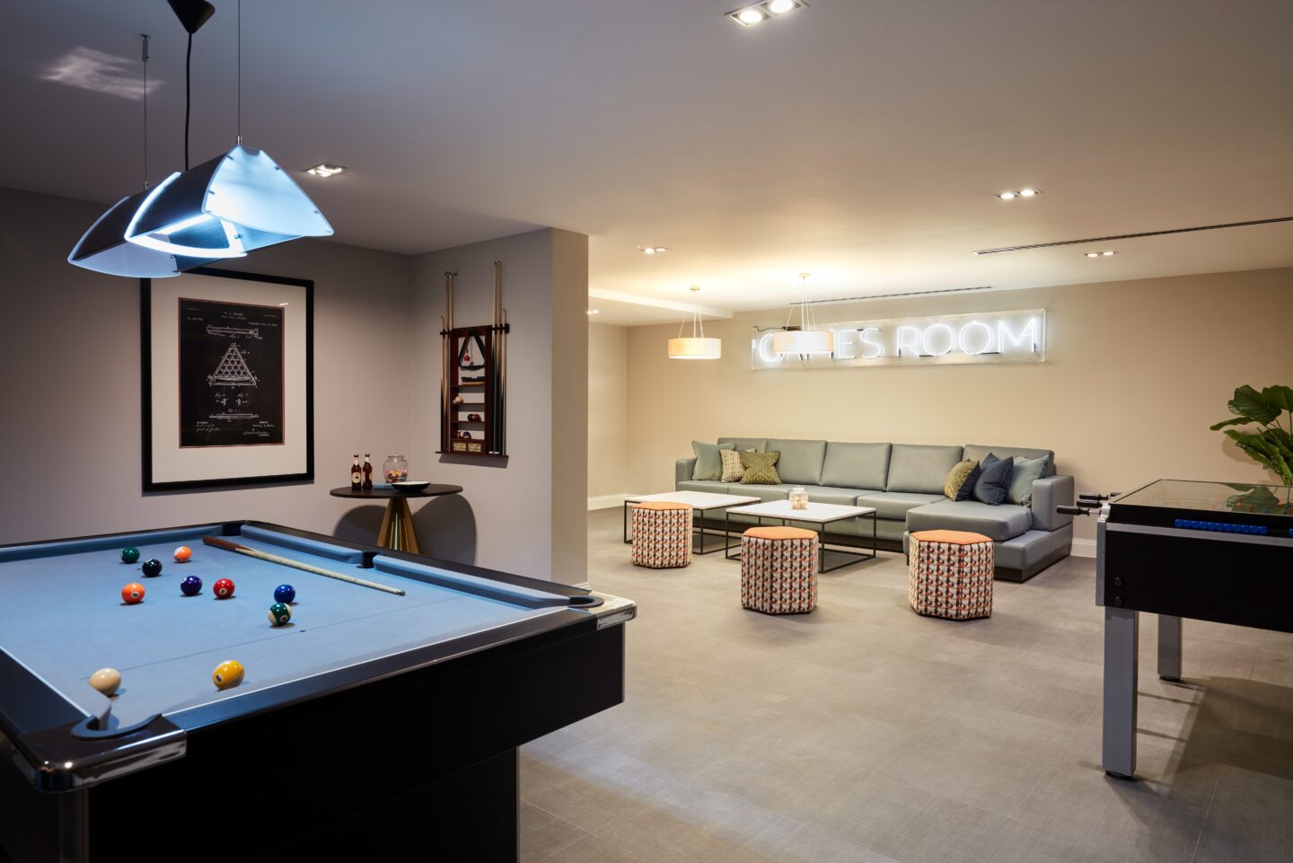 Modern games room with a pool table, table football, sofa, round pouffes, wall decor, and a neon sign reading GAMES ROOM on the back wall.