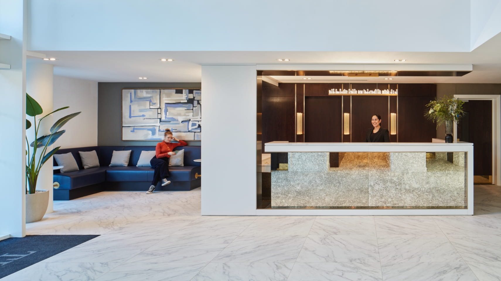 A modern hotel lobby features a reception desk with a smiling attendant, a seating area with a person sitting, abstract wall art, and marble flooring.