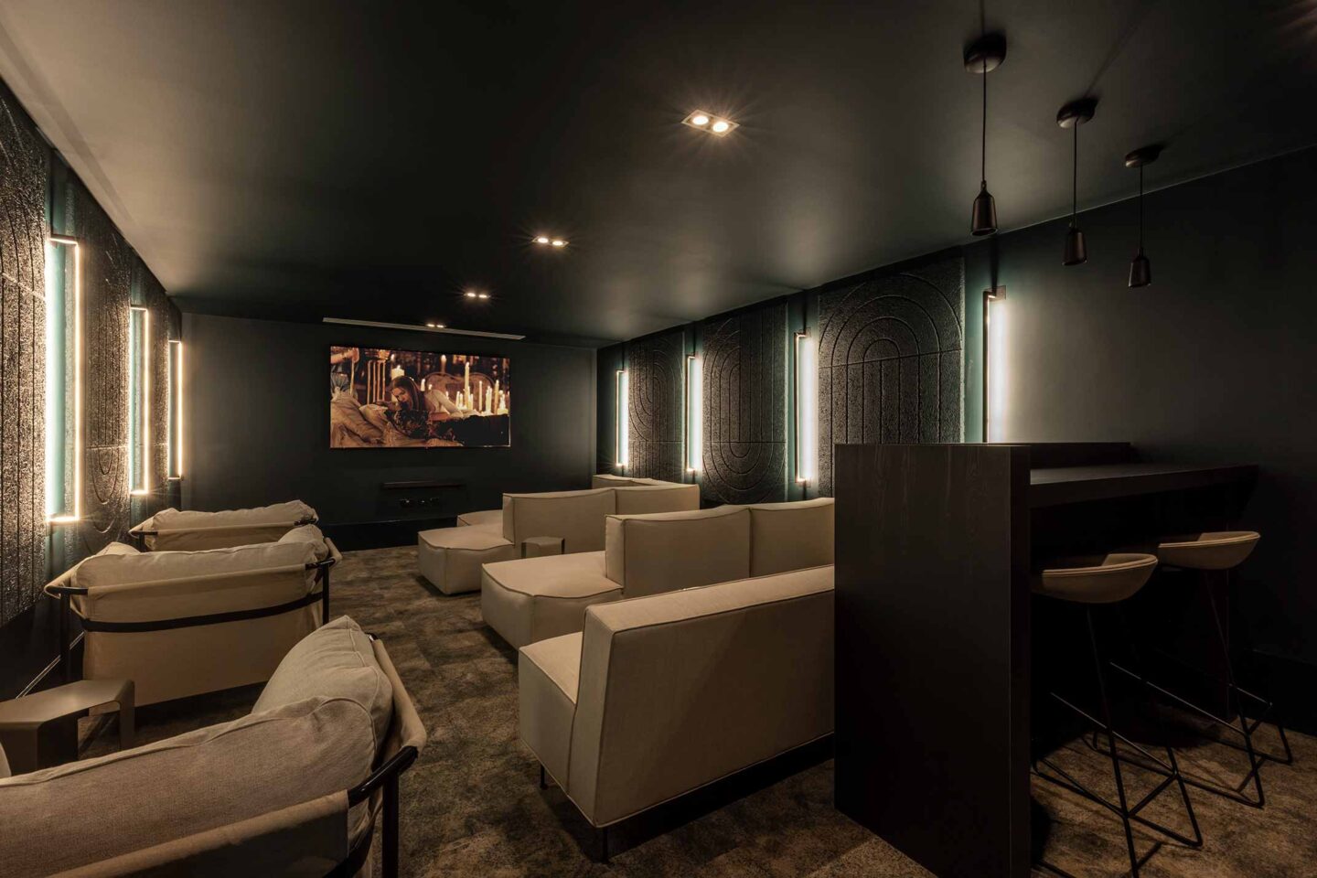 A modern home theatre room with beige sofas, wall-mounted lights, a large screen displaying a film, and a bar counter with stools in a dimly lit setting.