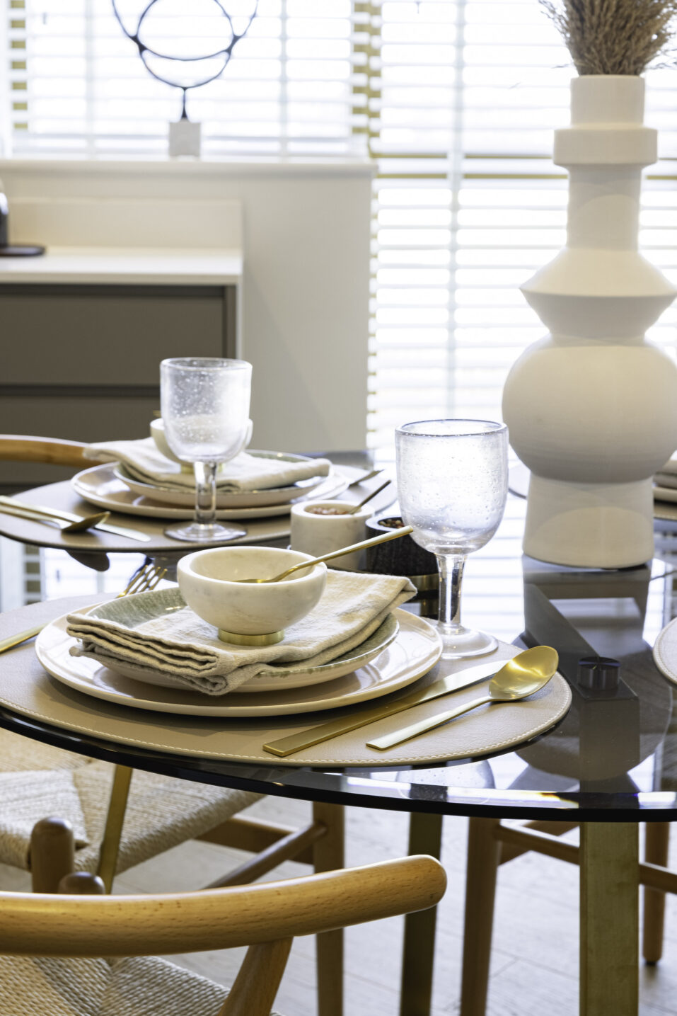 A glass dining table set with plates, bowls, serviettes, gold cutlery, and water glasses, with a white vase centrepiece and wooden chairs.