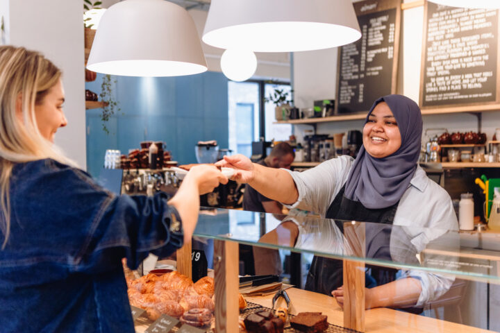 A customer hands a bank card to a smiling cashier behind a bakery counter with pastries and chalkboard menus in the background.