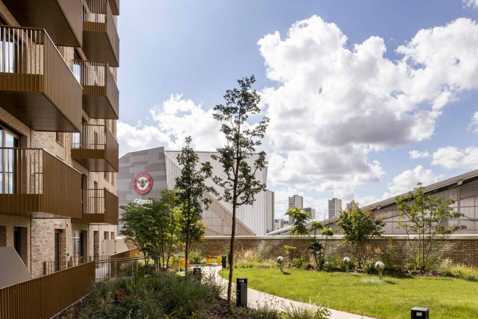 View of a landscaped courtyard with trees and grass, bordered by modern flat buildings and an adjacent stadium under a partly cloudy sky.
