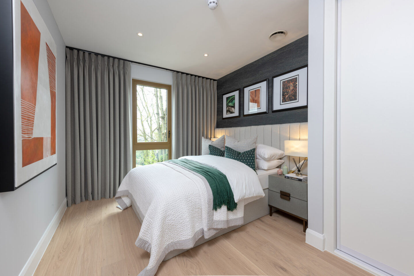 Modern bedroom with a double bed, white and green bedding, bedside tables, framed artwork above the headboard, large window with curtains, and light wood flooring.