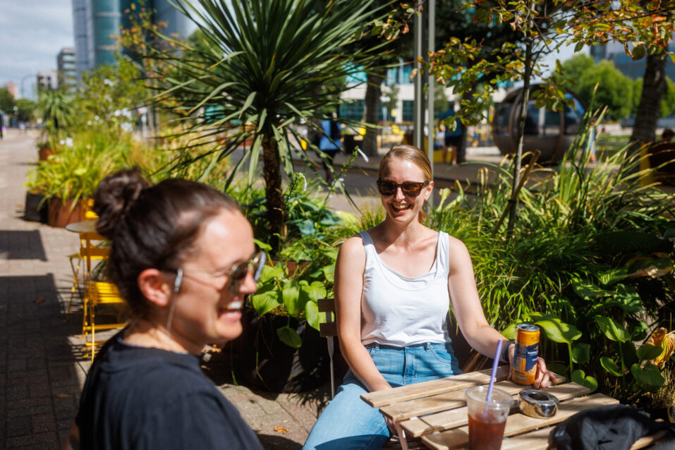 Two women sit at an outdoor table surrounded by plants on a sunny day. One drinks from a tin, the other from a cup with a straw. Both wear sunglasses and appear to be talking.