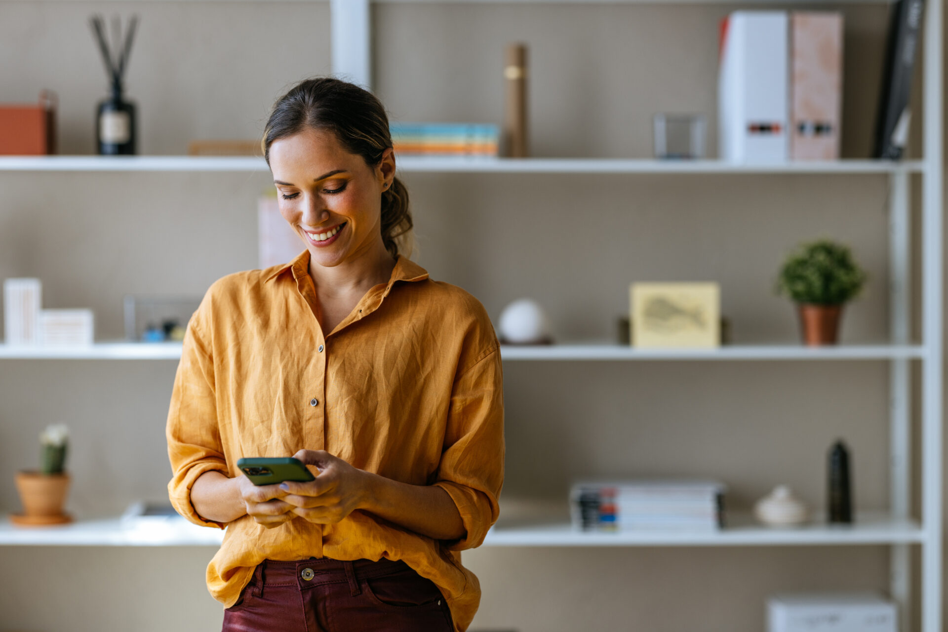 A woman in a mustard yellow shirt stands indoors, smiling whilst using her mobile phone. There are shelves with books and plants in the background.