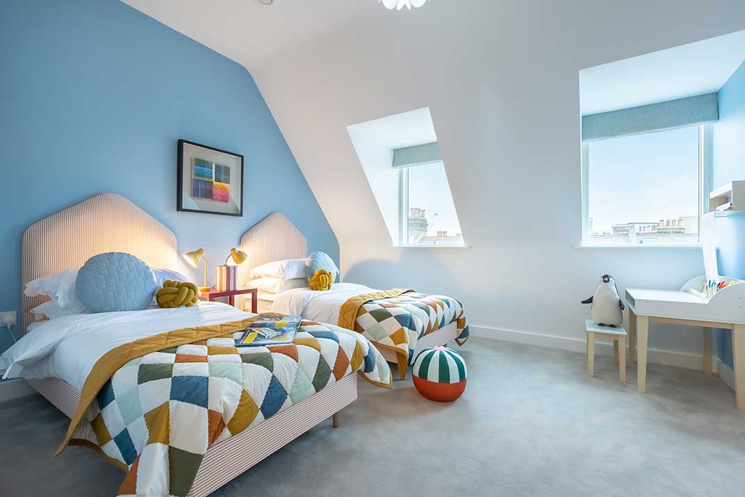 A bright bedroom with two single beds, colourful geometric quilts, and toys. The walls are partly blue, with two large windows and a small desk with a chair.