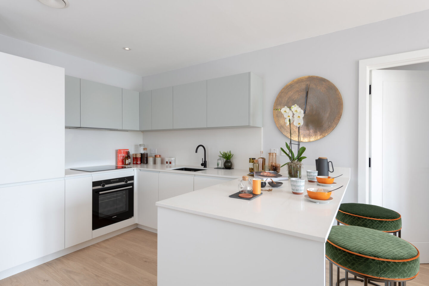 Modern white kitchen with built-in appliances, light grey cabinets, a black tap, green barstools, and a worktop set with dishes, glasses, and an orchid plant.