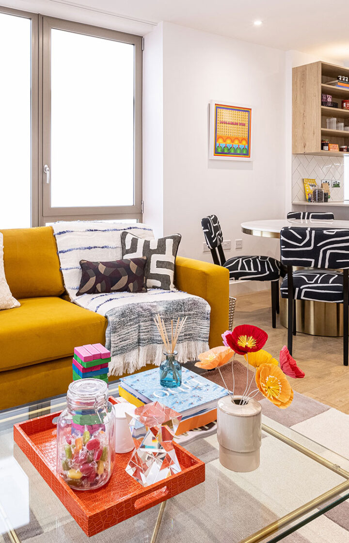 Modern living room with a mustard yellow sofa, geometric cushions, colourful decor items on a glass coffee table, and a dining area with patterned chairs in the background.
