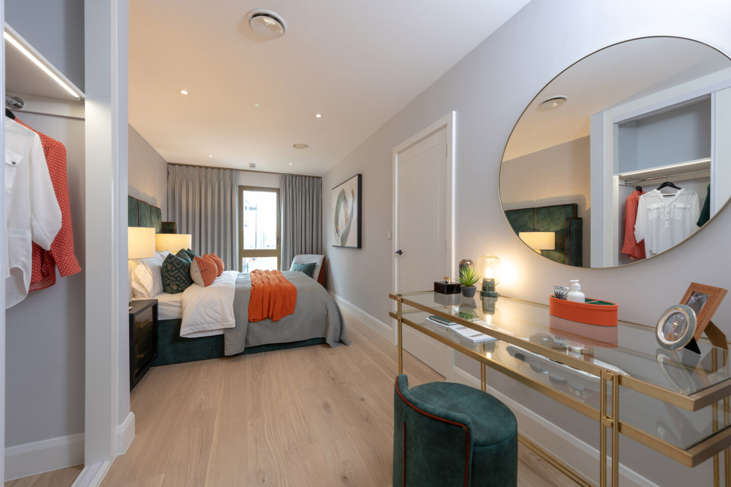 Modern bedroom with a bed, orange accents, fitted wardrobe, glass desk with a round mirror, and a window with grey curtains.