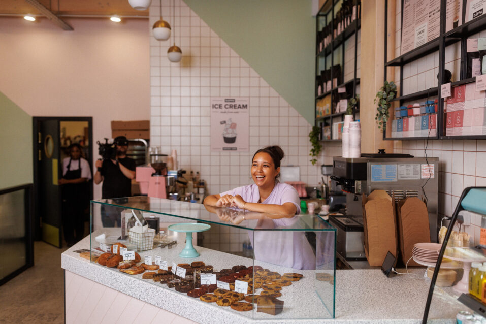 A woman stands behind a glass display counter with assorted pastries in a modern bakery. Shelves with supplies and a menu are visible on the wall behind her.