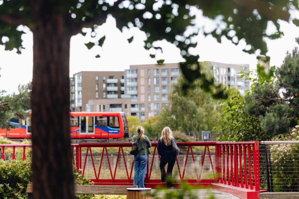 Two people stand on a red bridge, looking towards a block of flats and a passing red train, surrounded by trees and greenery.