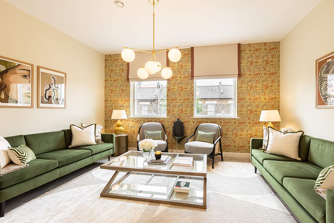 Bright living room with green sofas, glass coffee table, two chairs, patterned wallpaper, large windows, framed art, and a modern chandelier.