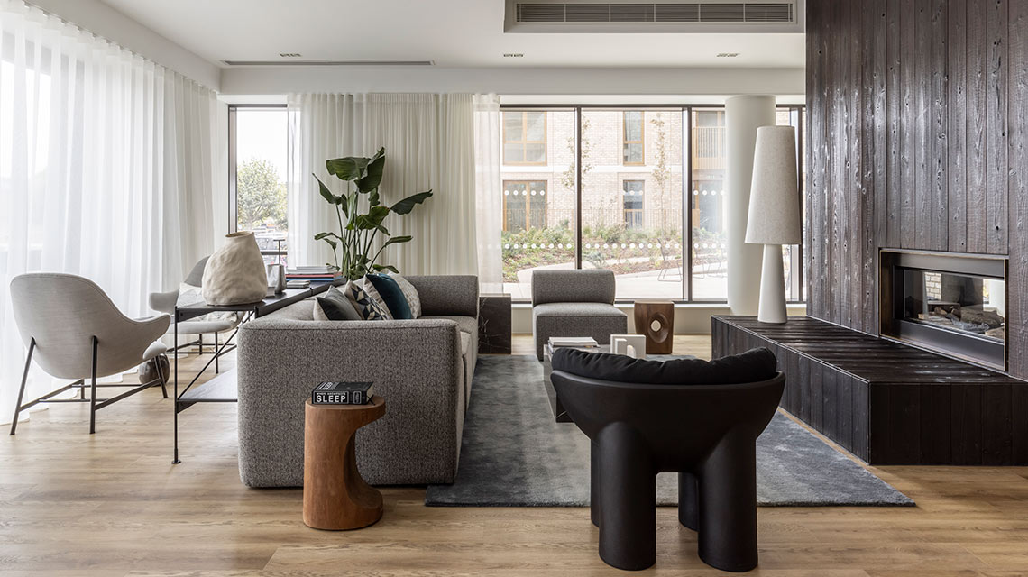 Modern living room with large windows, a grey sofa, contemporary chairs, a dark wood fireplace, and minimalist decor on a light wood floor.