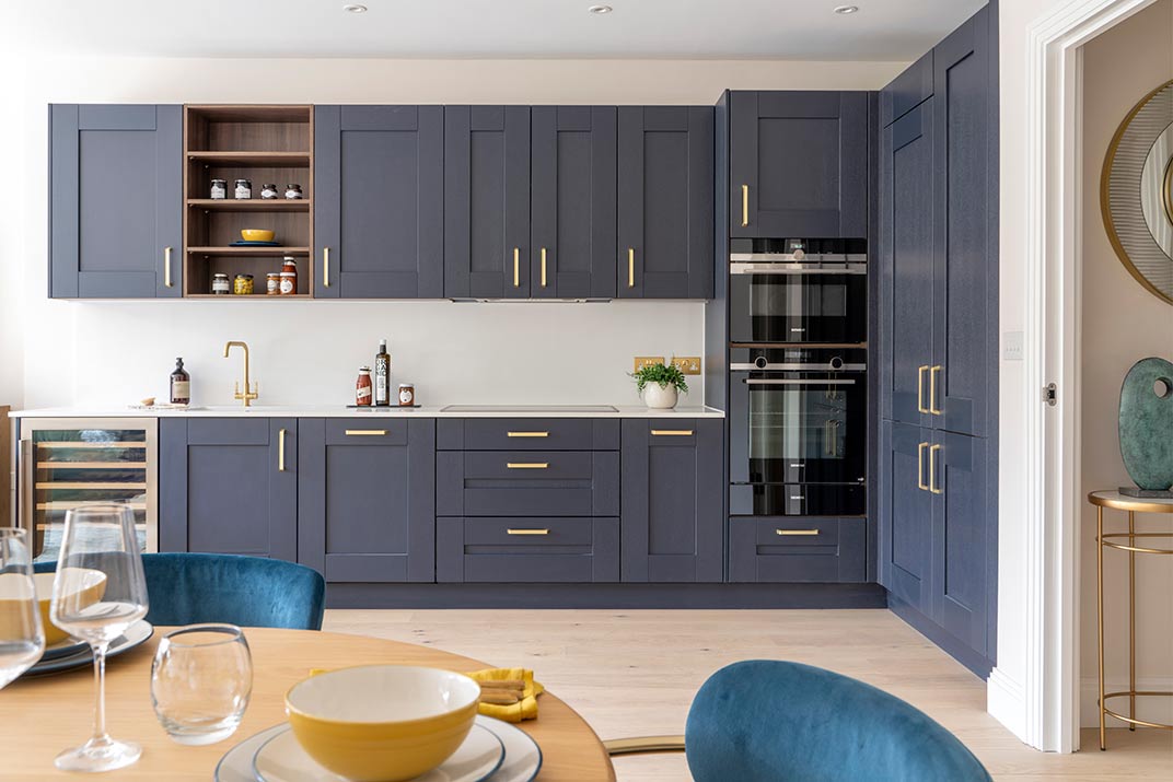 Modern kitchen with dark blue cupboards, gold handles, built-in double oven, open shelving, and a set dining table in the foreground.
