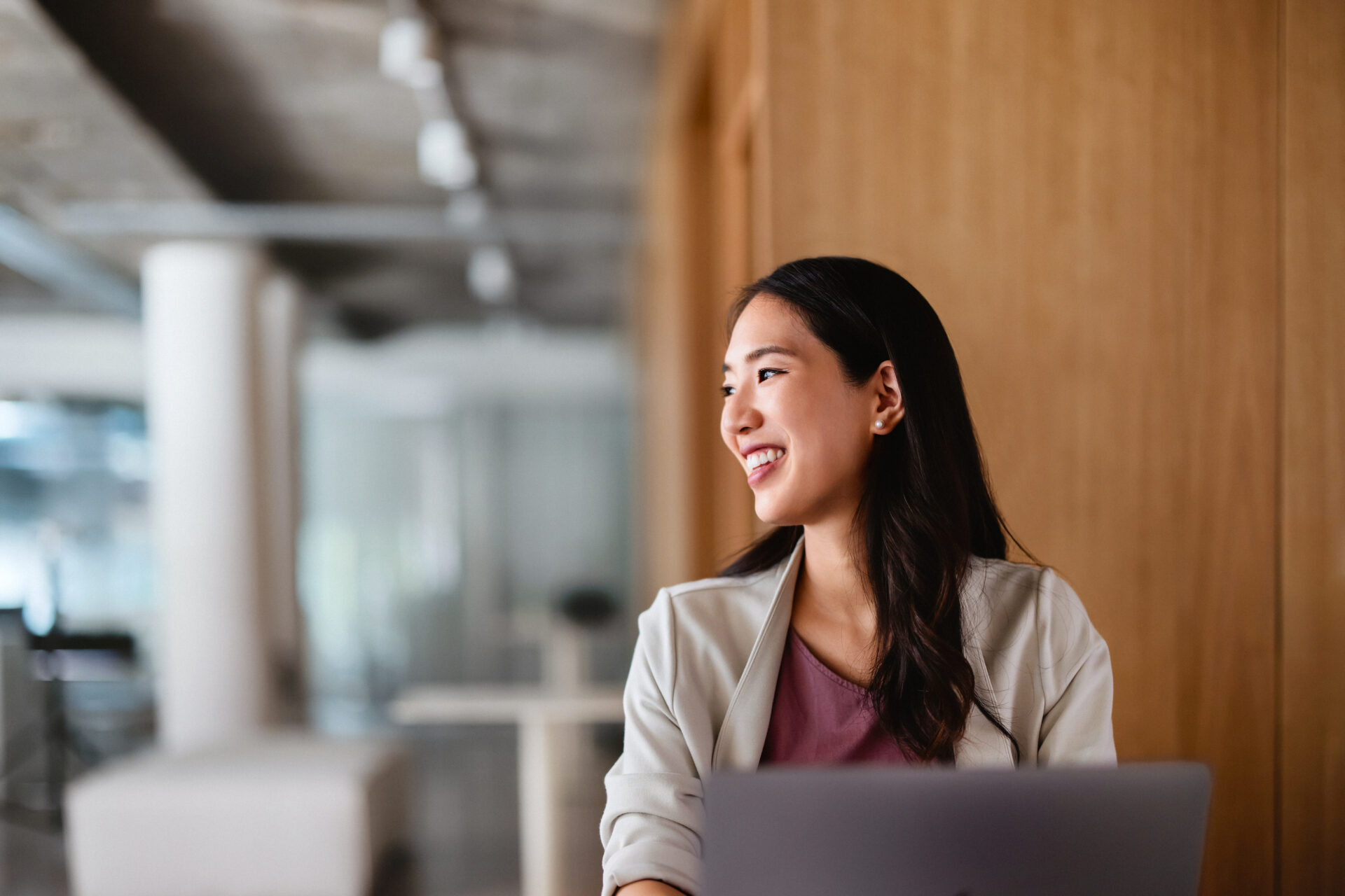 A woman sits indoors at a table with a laptop, smiling and looking to the side. The background features a wooden wall and a modern office setting.
