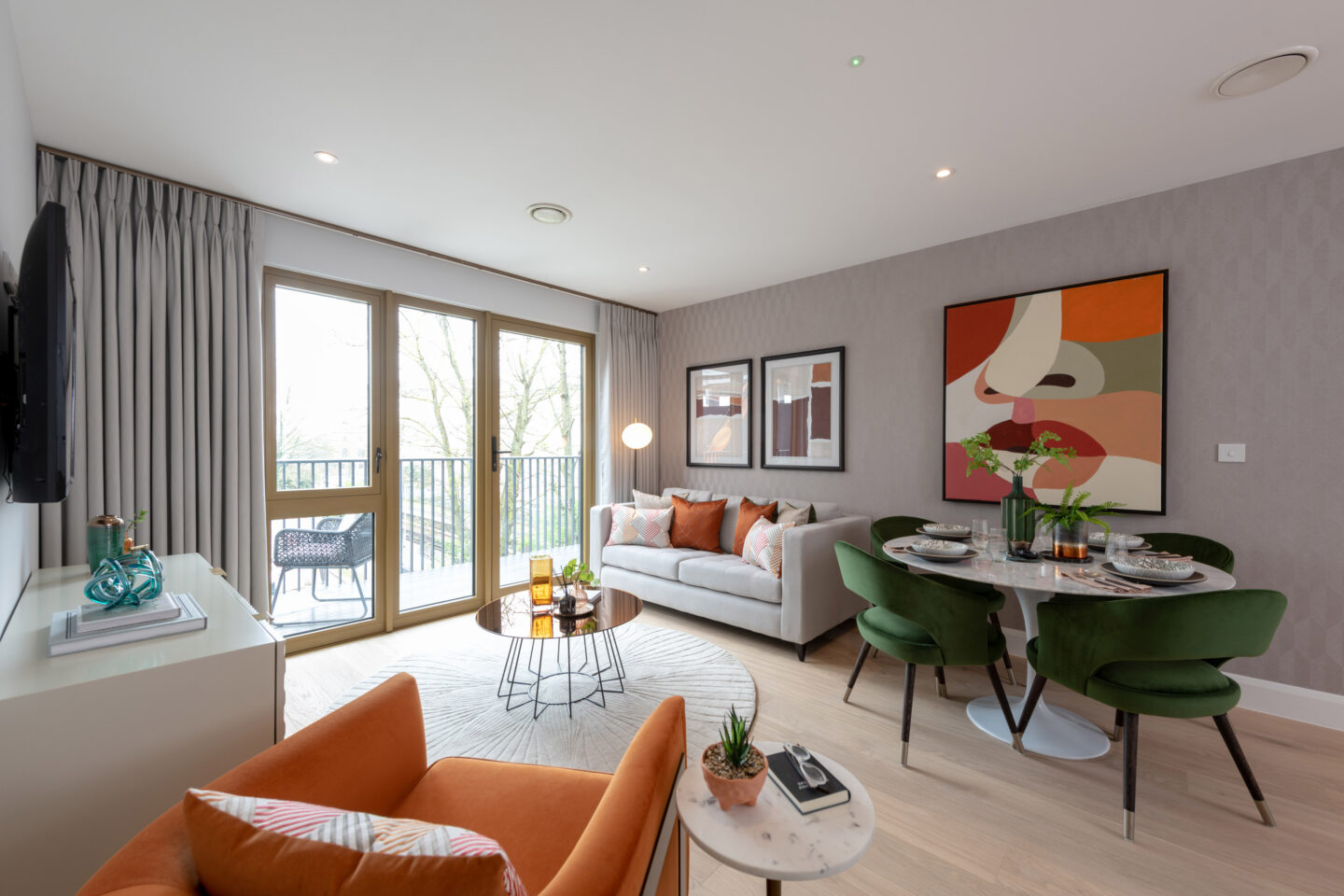 Modern living and dining area with a grey sofa, green dining chairs, orange armchair, large window, and abstract wall art. A round dining table is set for four.
