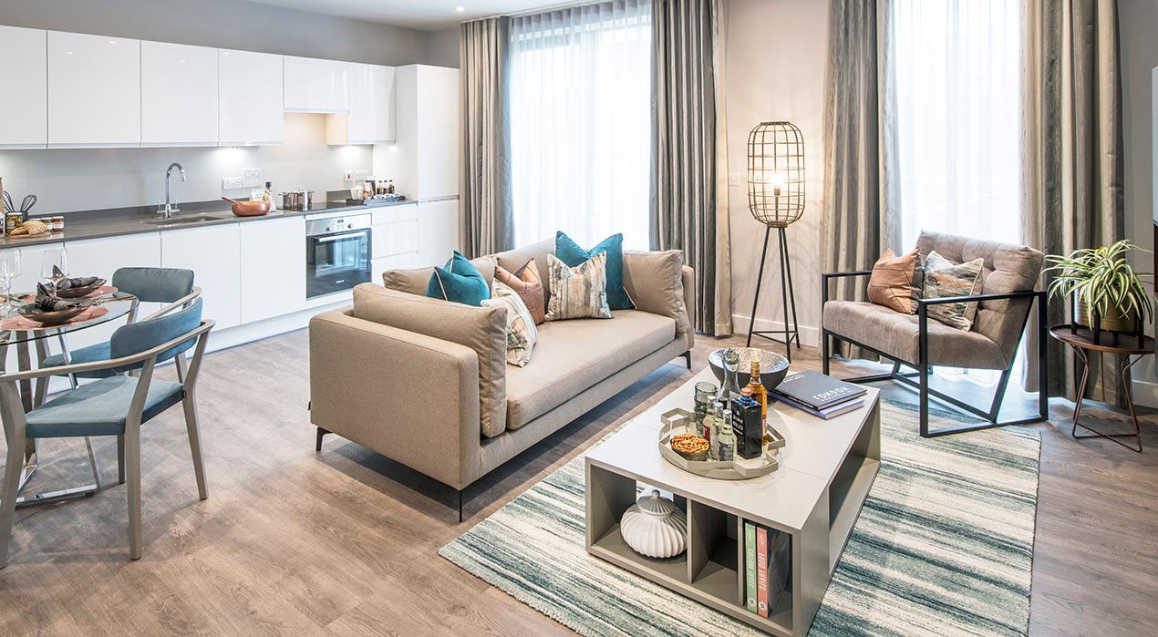 Modern open-plan living room and kitchen with neutral colours, a sofa, armchair, dining table, and floor-to-ceiling windows with sheer curtains, decorated with plants and books.