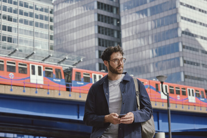 A man with glasses and a rucksack stands outdoors using his mobile phone, with a red train and tall office buildings in the background.