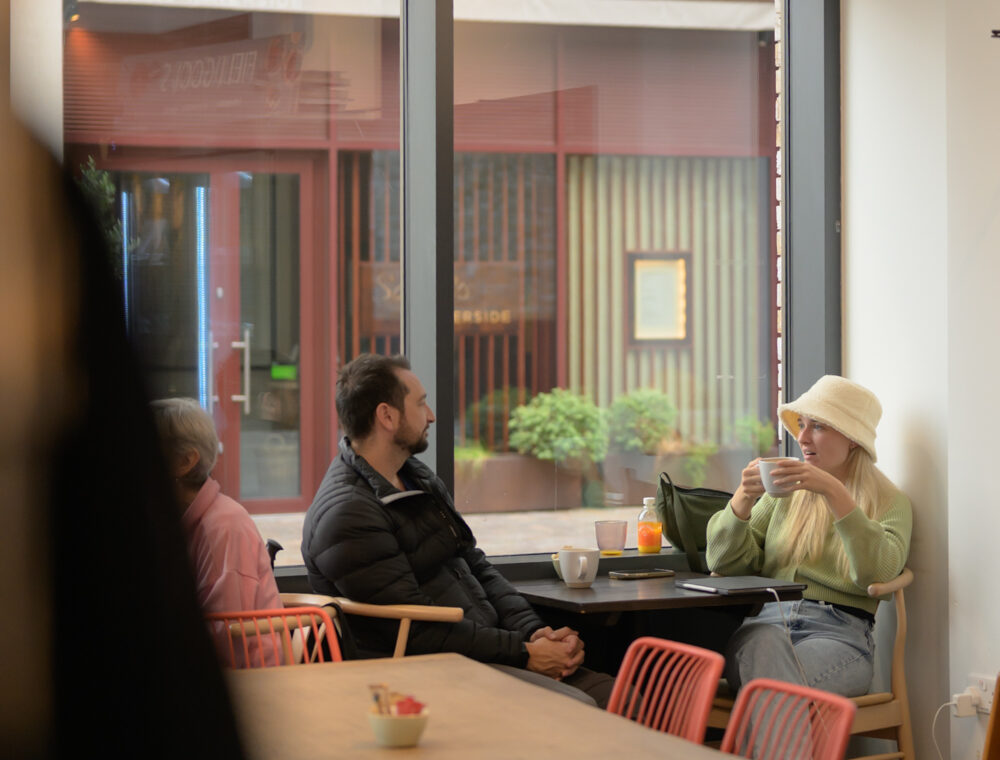 Three people sit at a table by a large window in a café; one woman in a light hat and jumper drinks from a cup while two others look on. Spherical ceiling lights hang above.