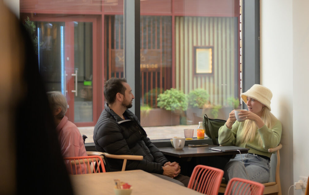 Two people sit at a table in a café near a large window; one person drinks from a mug, while the other looks in their direction. Spherical lights hang from the ceiling above.