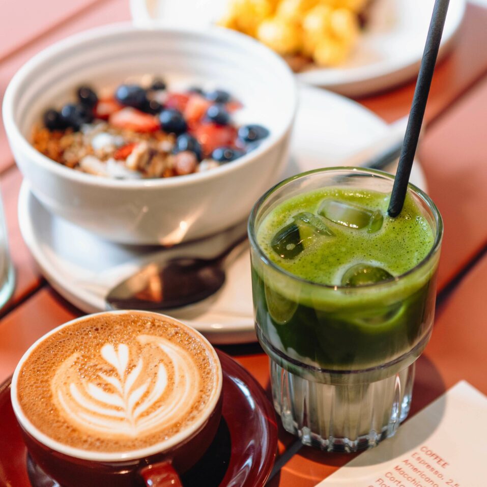 A latte with foam art, a glass of iced green juice with a straw, and a bowl of granola with fruit on a table.